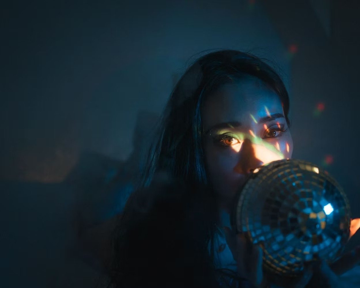 A person with long dark hair and makeup holding a disco ball close to their face, with colorful reflections on their face in a dark room. shot by Tokyo portrait photographer Deniz Demir.