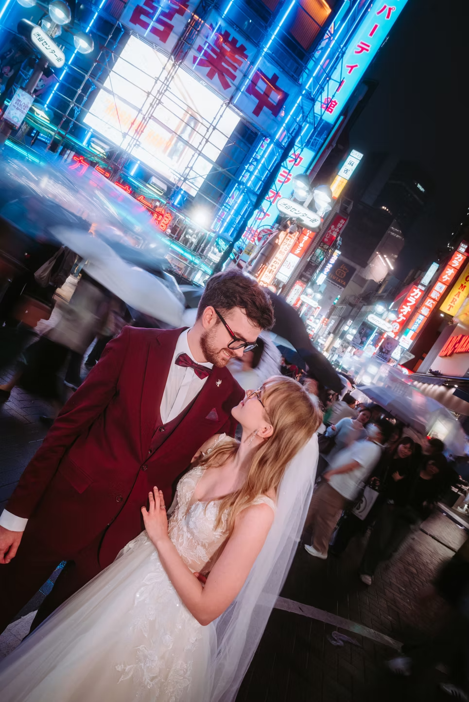 A couple dressed in wedding attire standing close in a busy city street at night, with neon signs and blurred pedestrians around them.