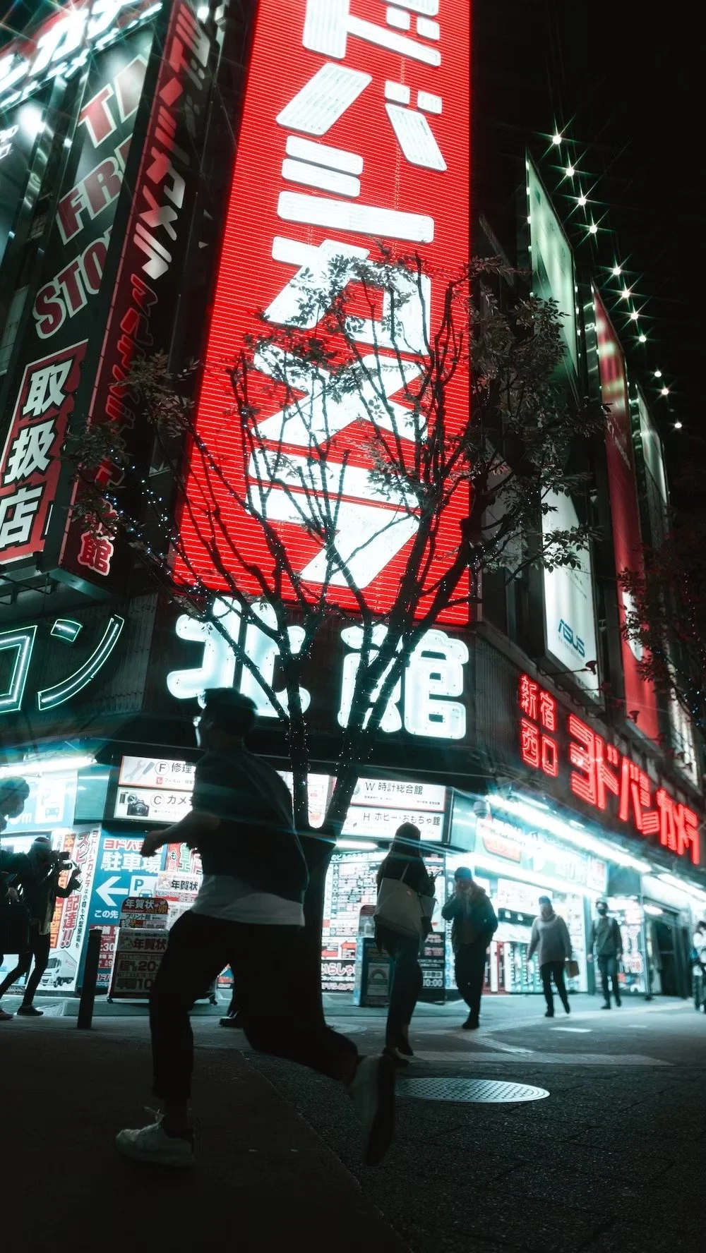 Nighttime city street scene with illuminated Japanese signs in Shinjuku, silhouette of a person in front of a tree, and people walking on the sidewalk.