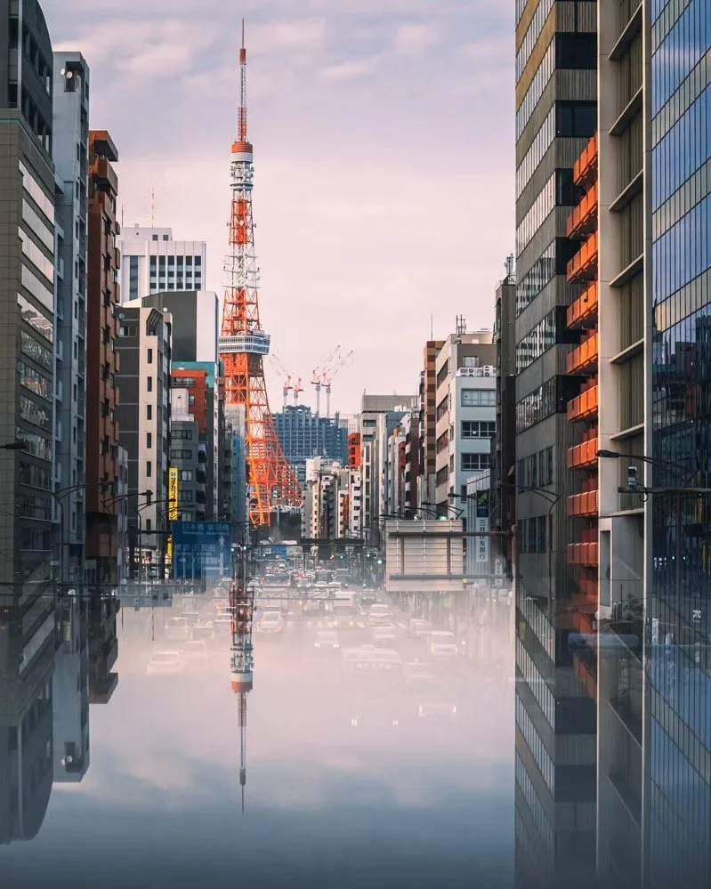 City street during daytime with tall buildings on both sides, Tokyo Tower visible in the background, and a reflection of the scene on water at street level.