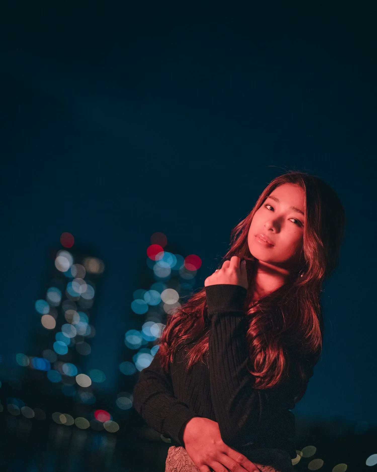 A young woman with long wavy hair poses against a dark background with colorful bokeh lights, wearing a black sweater and holding her glasses in her hand.