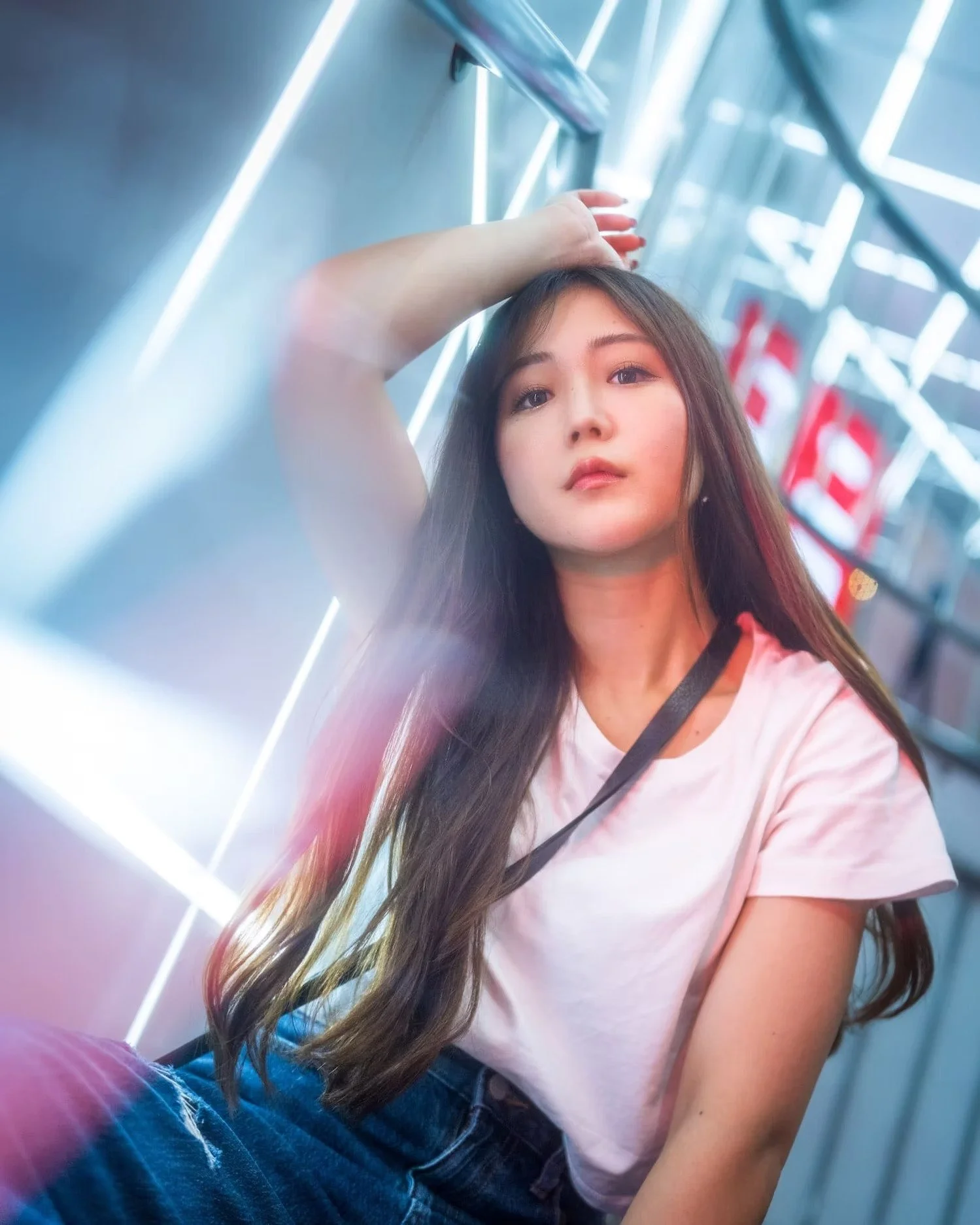 A young woman with long dark hair poses in front of neon lighting, with one hand resting on her head, inside a modern, illuminated space.