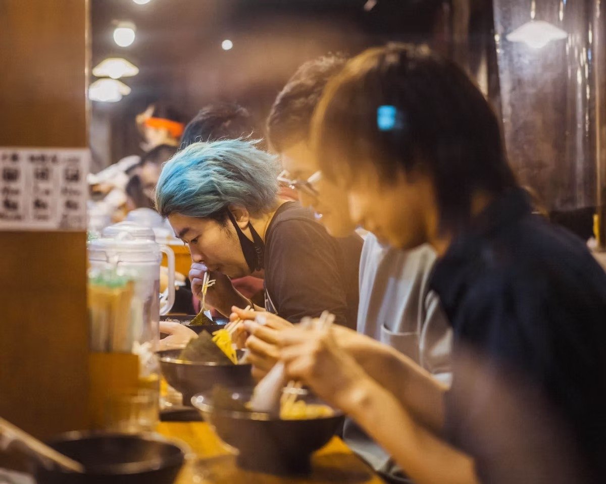 People eating ramen at a restaurant, focused on their bowls with chopsticks, in a busy dining area.