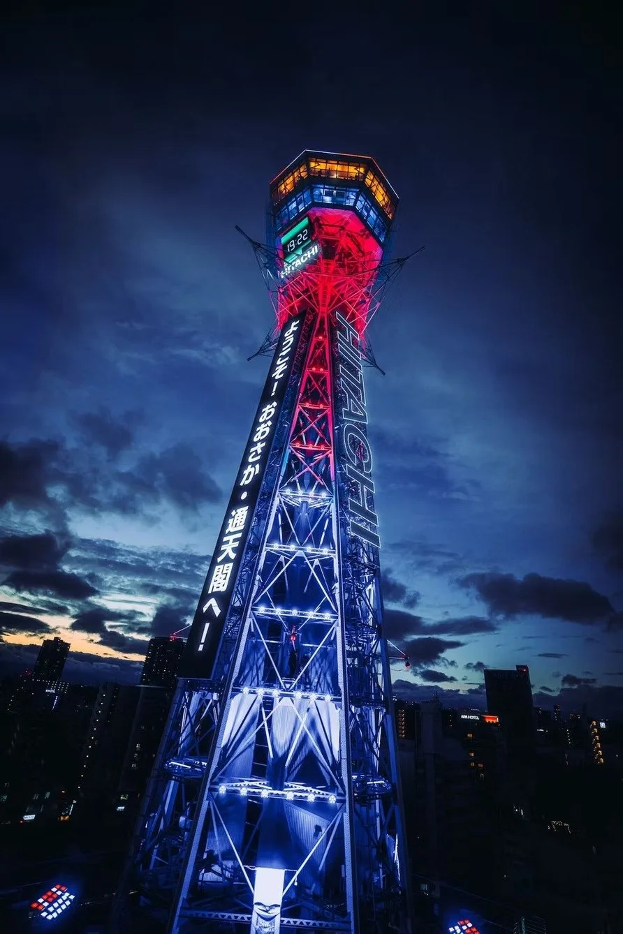 Night view of a tall Tsutenkaku tower in Shinsekai Osaka illuminated with red, blue, and yellow lights, with Japanese text on an electronic display board and a city skyline in the background. Shot by Deniz Demir, Tokyo photographer.