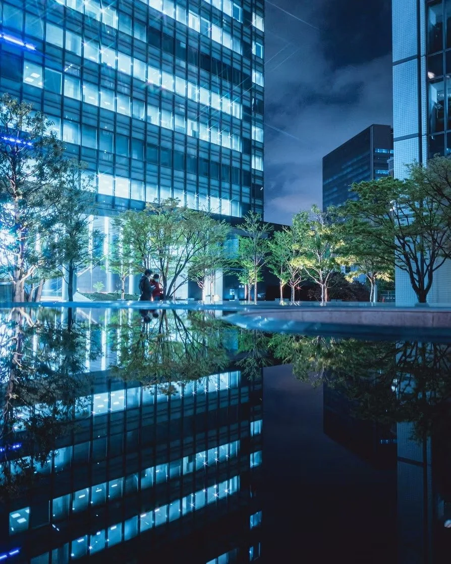 Nighttime cityscape with tall glass buildings, illuminated windows, and a reflective pool or water feature with trees.