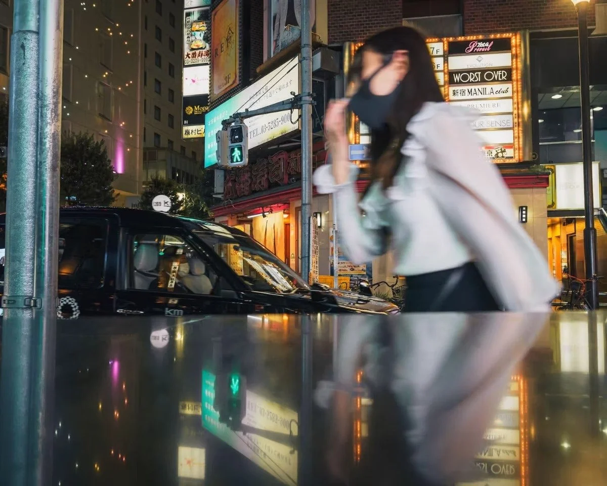 A hostess wearing a white jacket and black face mask standing on a city street at night, surrounded by bright neon signs and a car parked nearby.