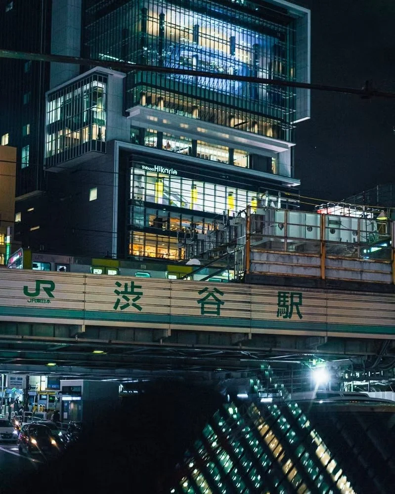 Nighttime view of Shibuya Station in Tokyo, Japan, with illuminated modern buildings and traffic below.