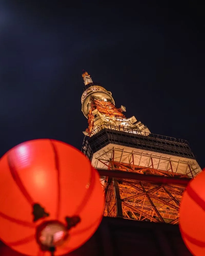 Night view of Tokyo Tower illuminated with orange lights, seen behind red paper lanterns.