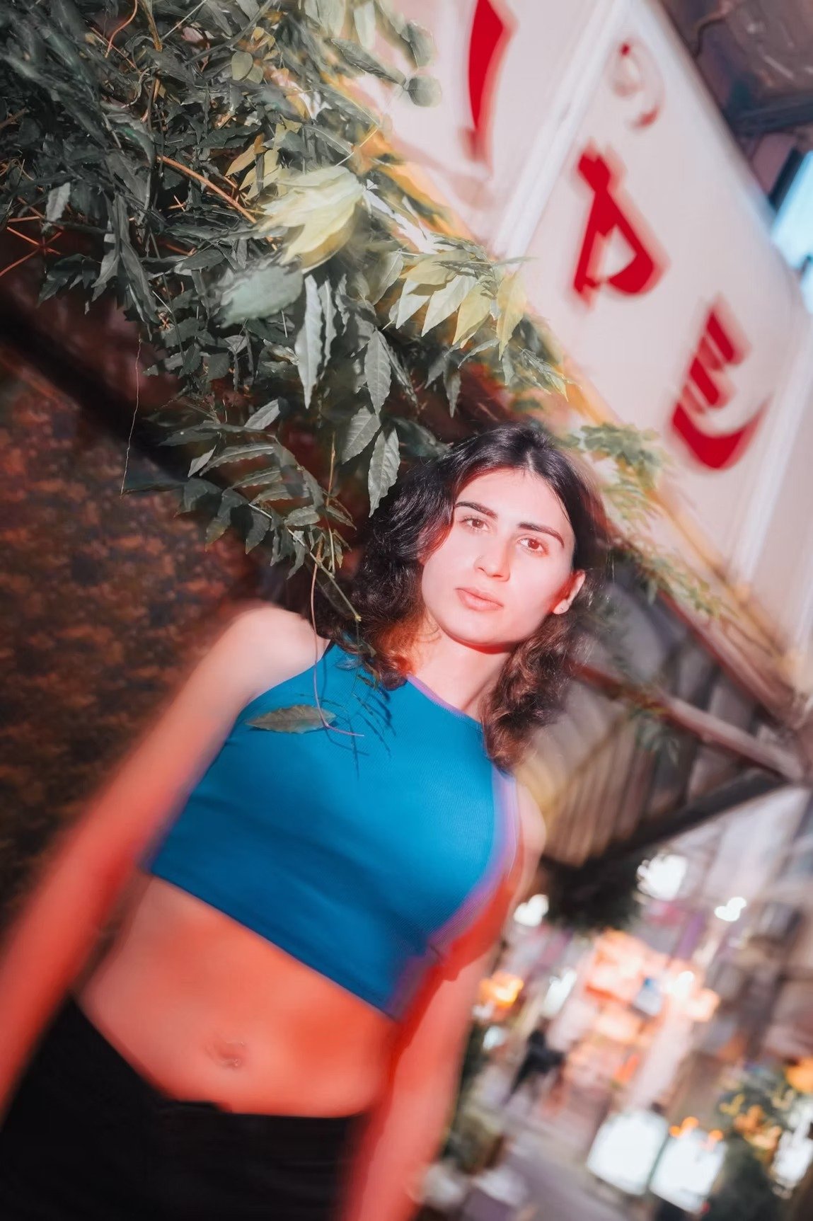 A woman poses in front of a vibrant city backdrop, wearing a casual cropped top. The motion blur effect captures the energetic atmosphere of the city, highlighting her effortless street style.