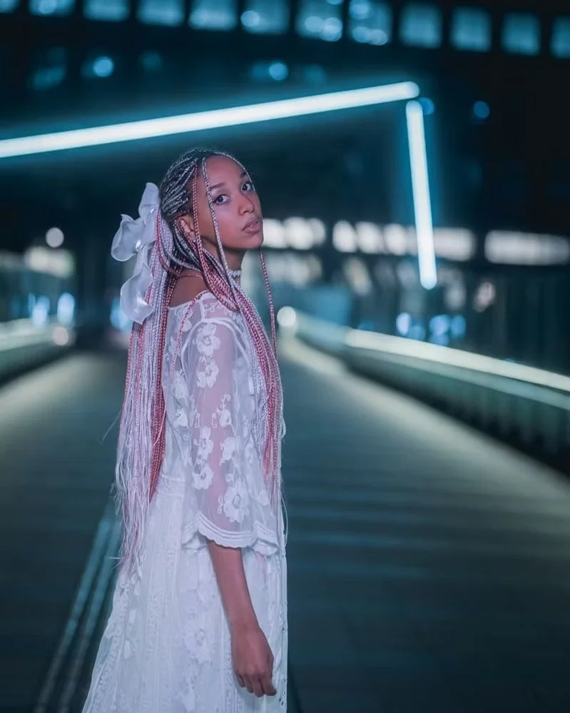Young woman with pink and white braided hair wearing a white embroidered dress standing on a modern illuminated bridge at night.