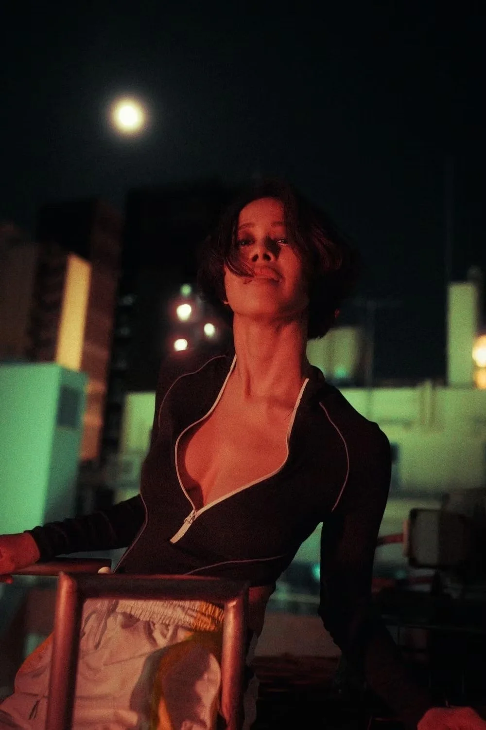 A woman with short dark hair, wearing a black zip-up top, stands on a Tokyo rooftop at night with a bright full moon overhead. The city lights are blurred in the background.