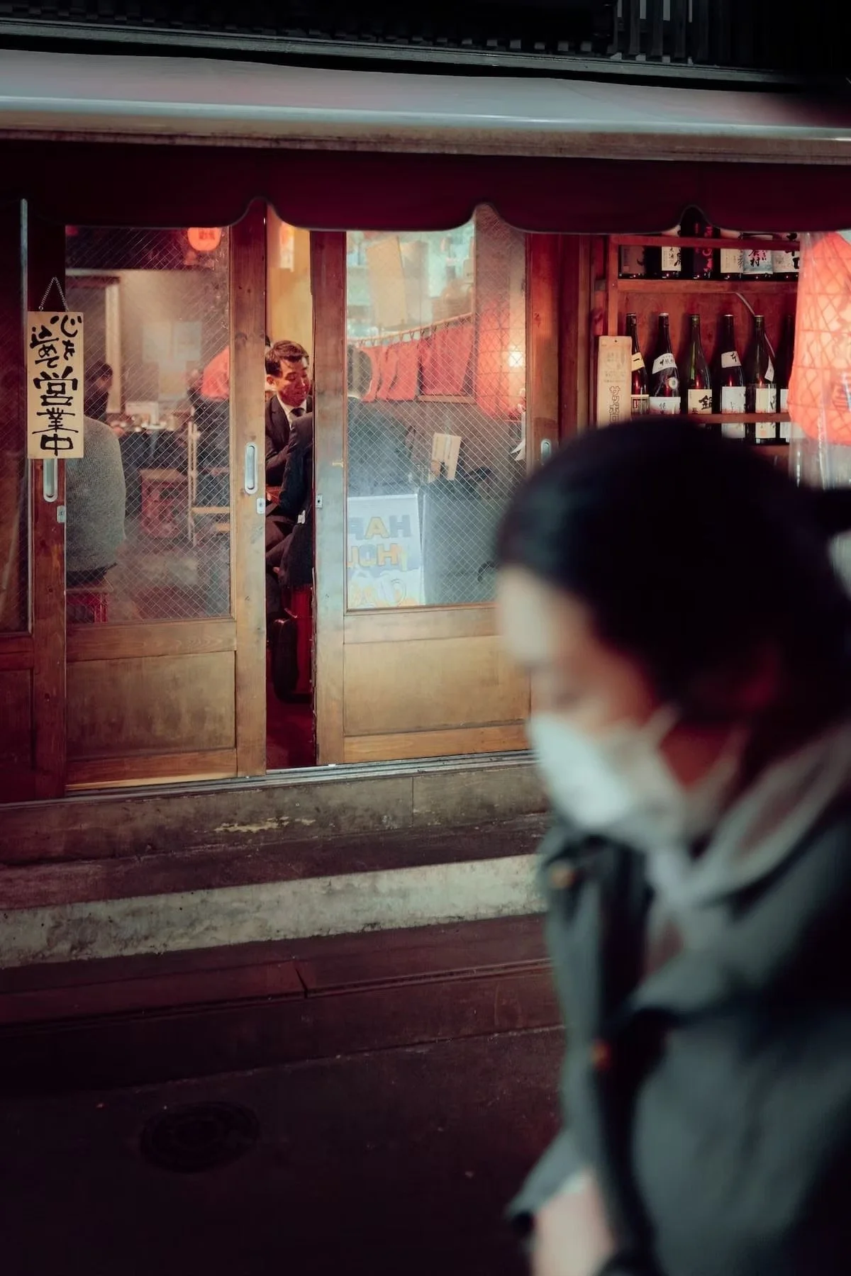 A woman wearing a face mask walking past a Japanese restaurant with a glass door, inside there are seated diners, and shelves with sake bottles are visible. Shot by Tokyo street photographer Deniz Demir.
