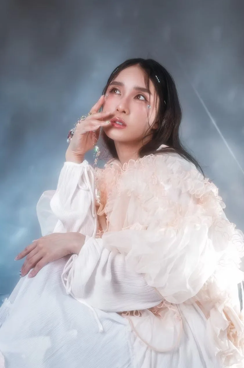A woman dressed in a flowing white outfit poses gracefully against a dreamy, soft-lit background. Her serene expression enhances the angelic and delicate atmosphere.