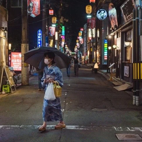 A woman in a blue kimono walks across a lantern-lit street at night, holding an umbrella and a shopping bag, surrounded by glowing signs and buildings.