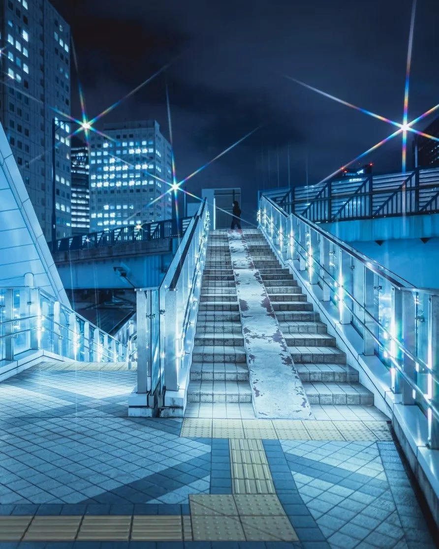 Night scene of a cityscape with illuminated buildings, a staircase with blue lighting, and a person standing at the top of the stairs on a rainy evening.
