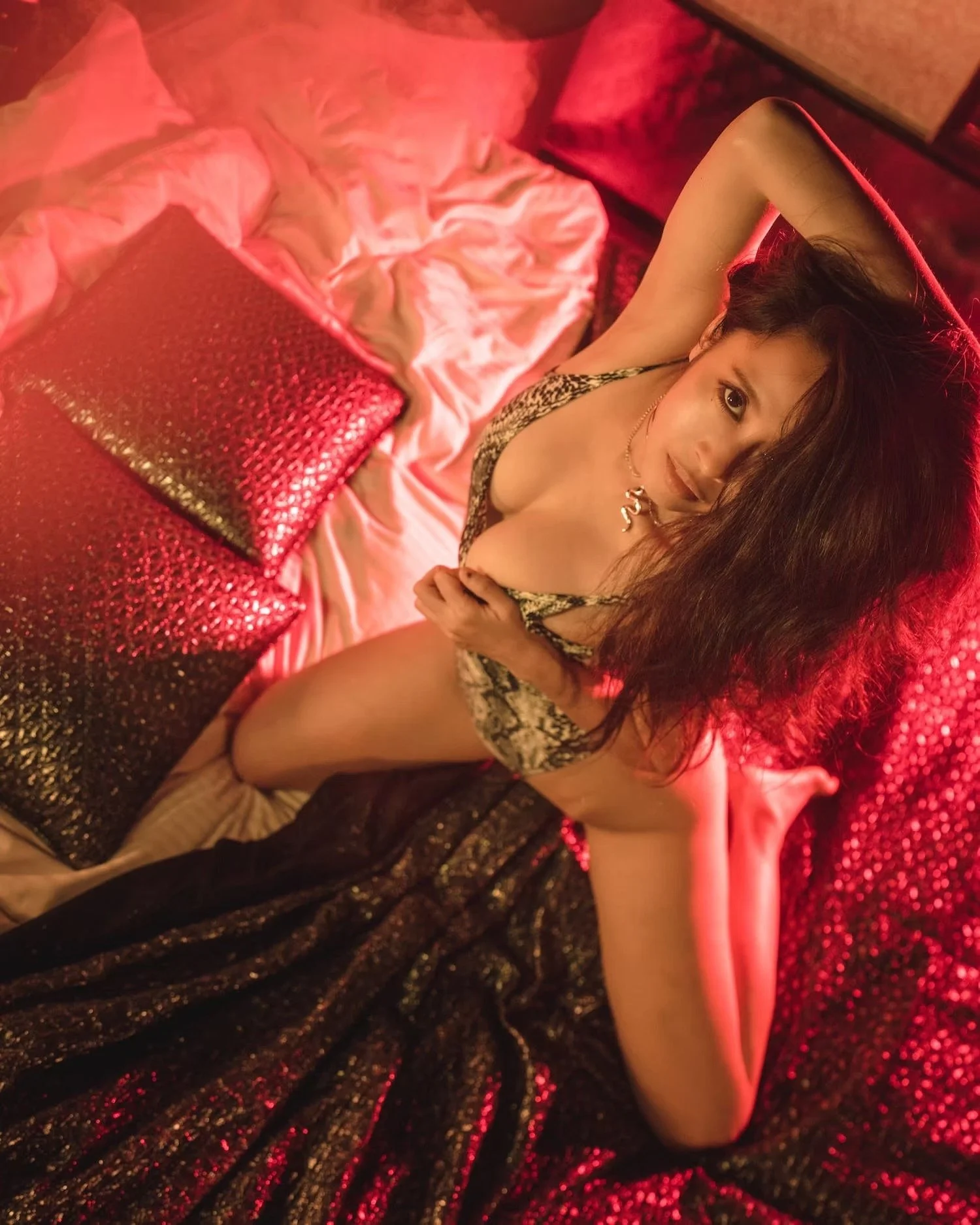 A woman lying on a bed with a satin sheet and a glittery pillow, wearing a leopard print dress and posing with her arm raised above her head, illuminated by red lighting.