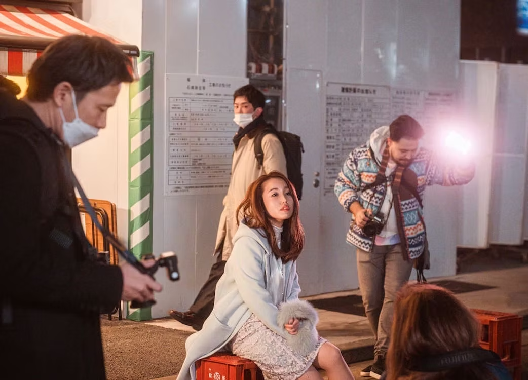 A woman sitting on a small red stool, surrounded by a group of people including photographers and videographers, at night outside a building.