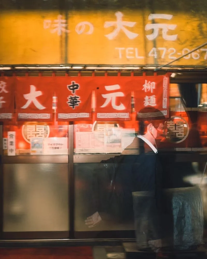 Reflected image of a person wearing a suit, facing right and wearing a face mask, standing in front of a storefront with red, yellow, and white signage in Japanese.