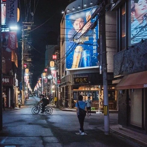 A woman walks down a dimly lit Tokyo street at night, surrounded by glowing billboards, bicycles, and hanging lanterns illuminating the scene.