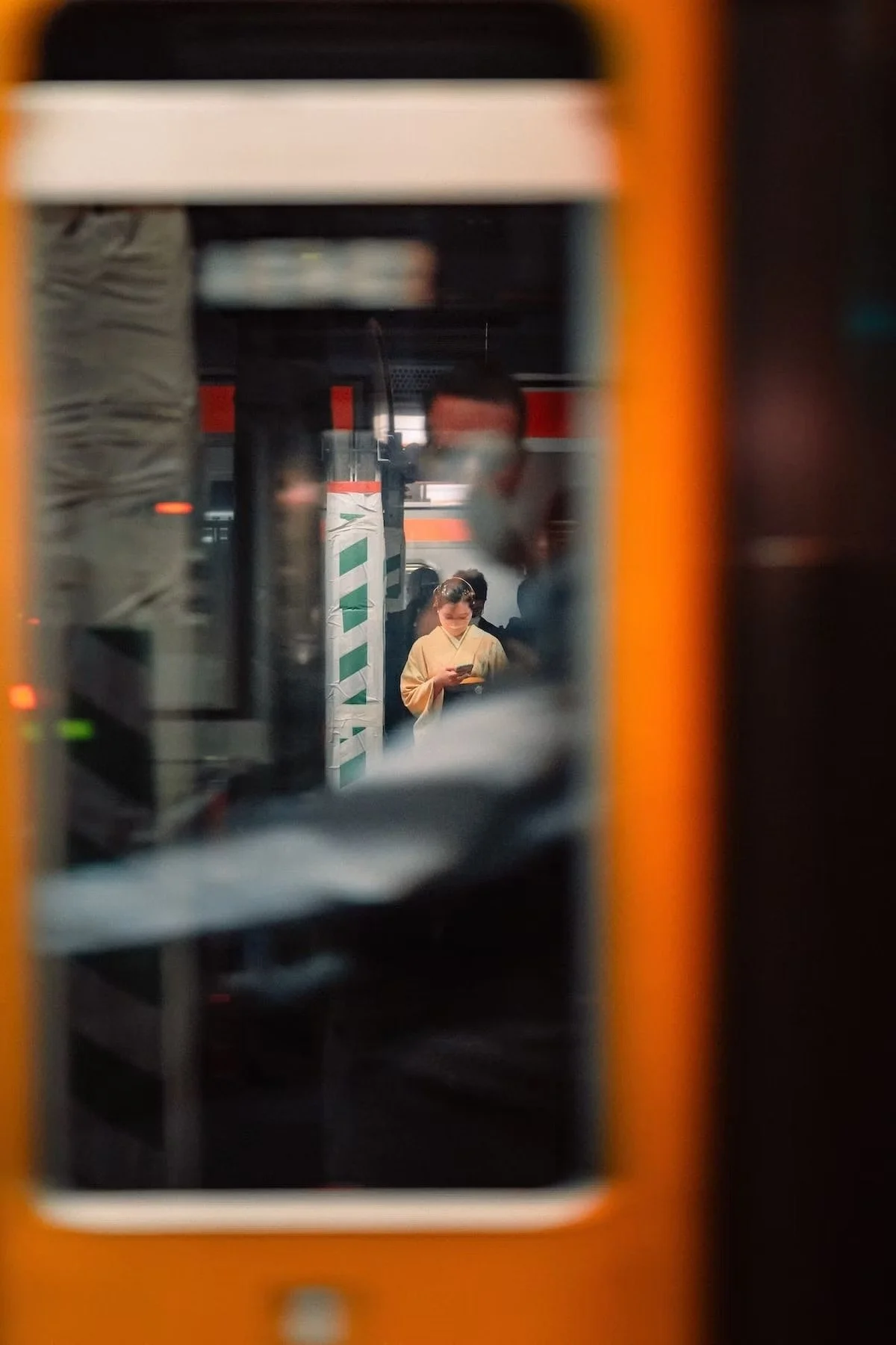 A Japanese person in a kimono looking at their phone among a crowd, seen through a narrow opening in a structure or frame with the train conductor in view and Deniz Demir in the reflection.