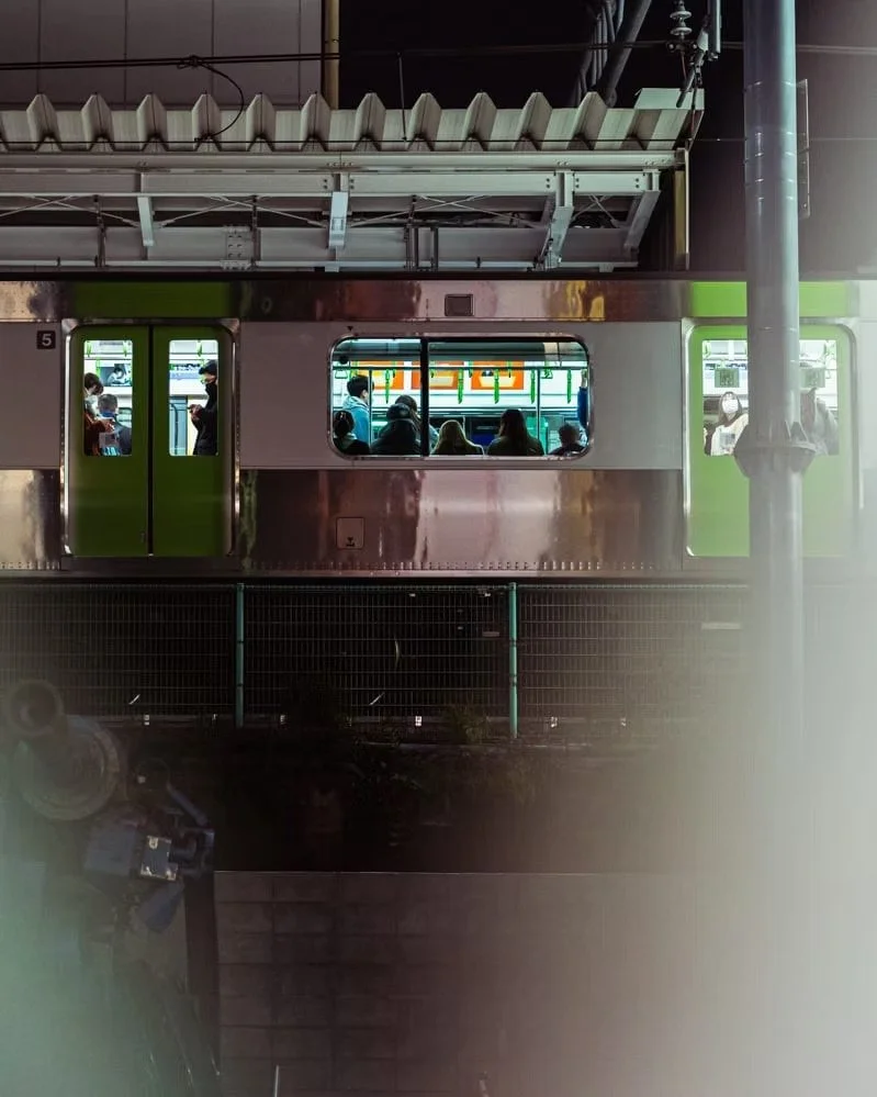People sitting and standing inside a train at a station, viewed through train windows, with a platform and a fence in front.