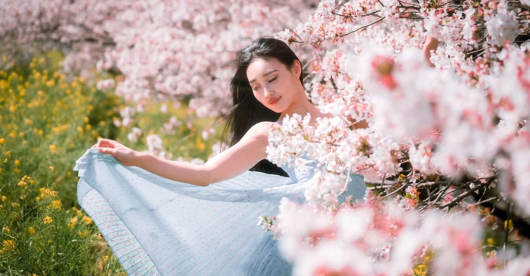 A woman in a white dress standing among blooming cherry blossom trees and yellow flowers, holding her dress and looking down with a gentle smile.