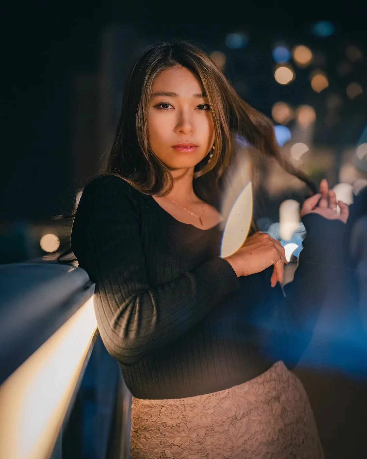 A woman sitting on a boat or ferry at night, with city lights in the background. She has long hair, is wearing a black long-sleeve top, a light-colored lace skirt, and a necklace. She is looking at the camera with a slight smile.