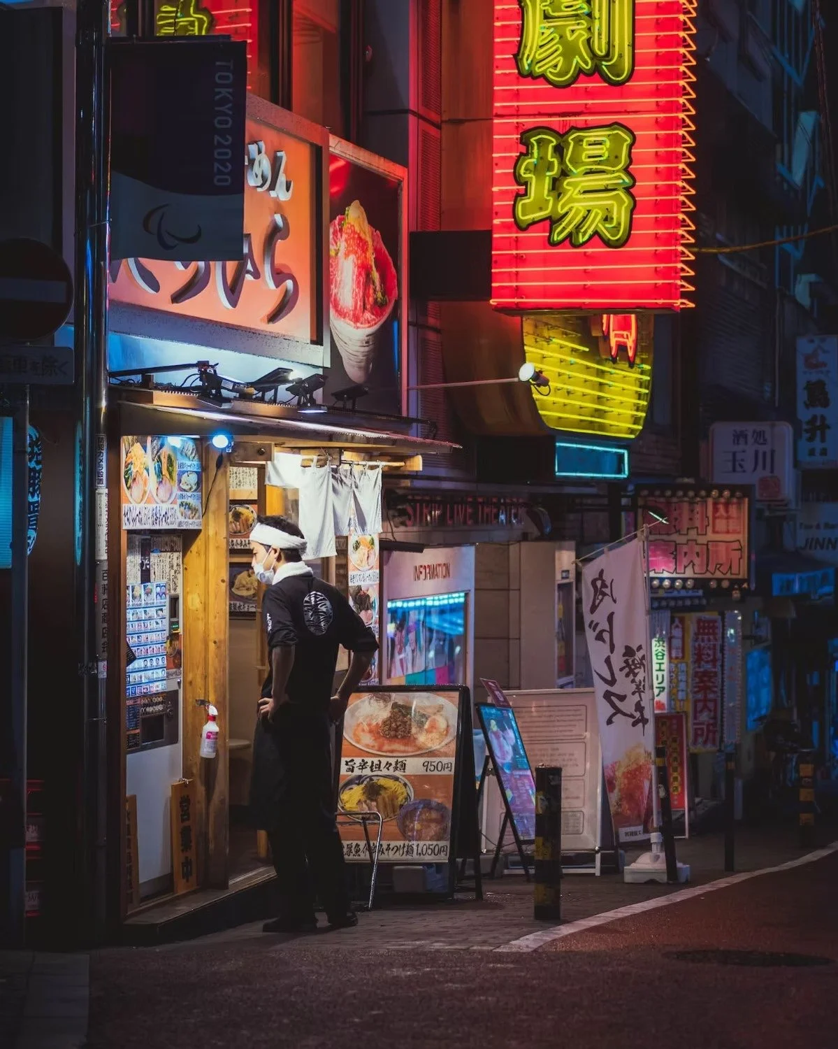 A nighttime street scene in Shibuya Tokyo Japan with colorful neon signs and a restaurant entrance. A person wearing a face mask and headband stands outside, looking into the restaurant. Neon signs in Japanese characters are visible, along with menu 