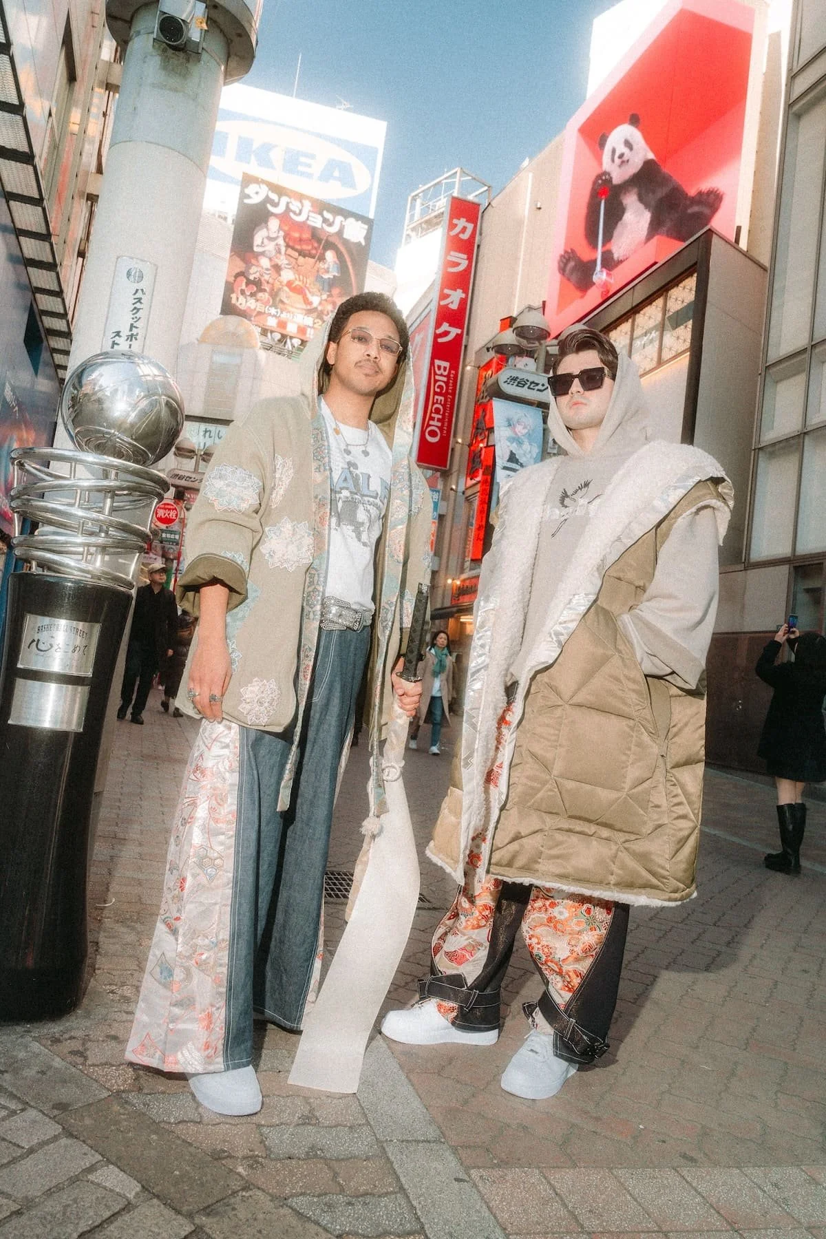 Two men dressed in fashionable streetwear standing on a busy city street with billboards and signs in the background. One man holds a sword, the other wears large sunglasses and unconventional clothing.