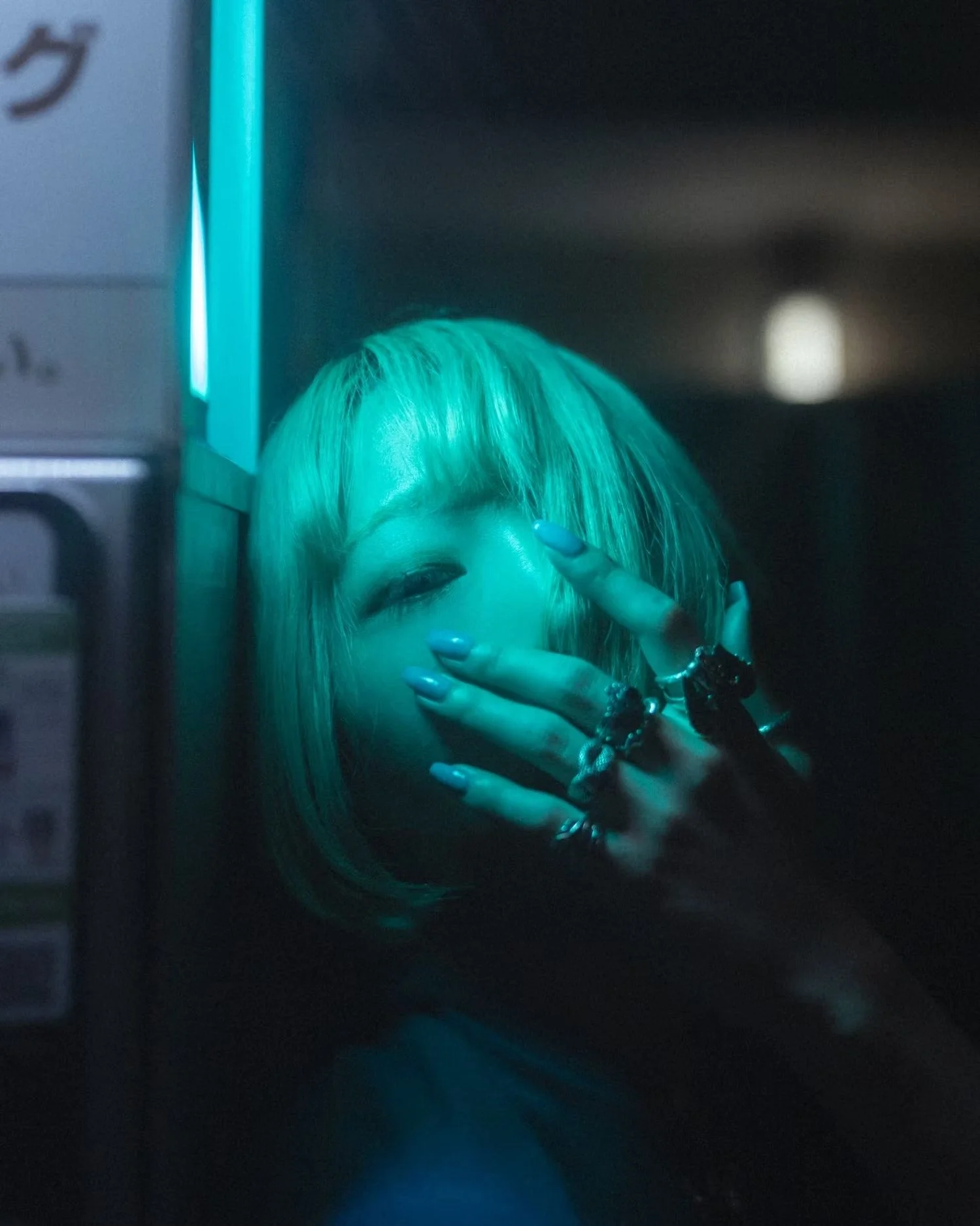 A person with teal-colored hair taking a mirror selfie, partially covering their face with their hand, in a dark setting with blue lighting.
