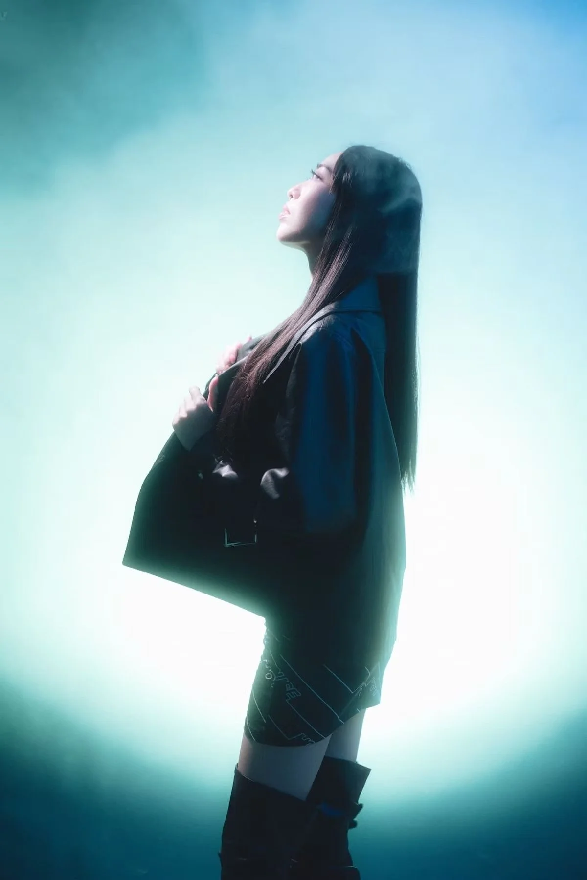 A young woman with long dark hair, wearing a black jacket and shorts, is standing against a greenish-blue gradient background. She is looking upward and holding a document or folder close to her chest. Studio portrait photography by Deniz Demir.