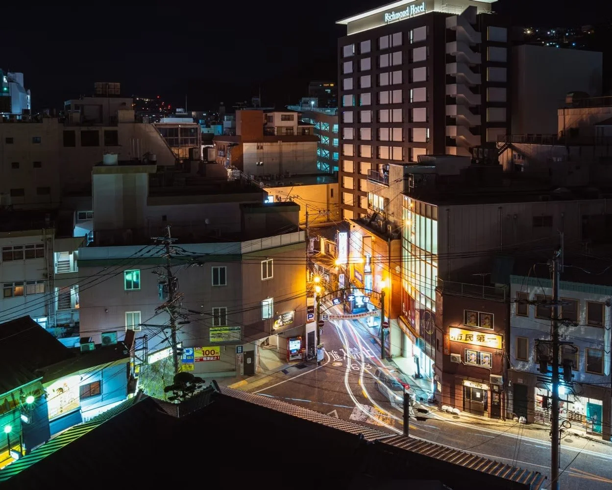 Nighttime city street scene with illuminated buildings, streetlights, and power lines. A tall hotel named 'Richmond Hotel' is visible in the background.