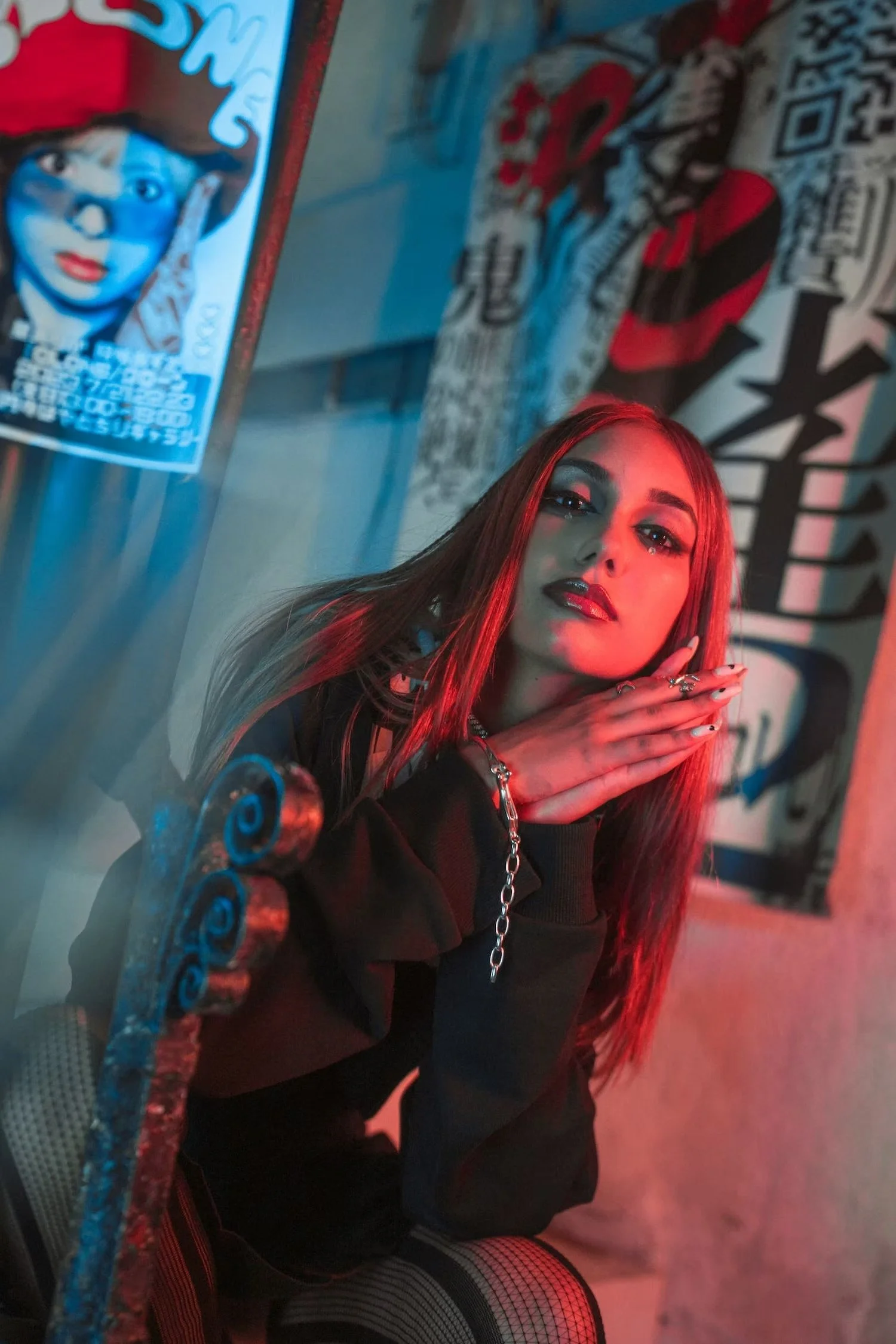 A young woman with long red hair and dark makeup posing indoors with graffiti art and posters in the background, illuminated by red and blue lighting.