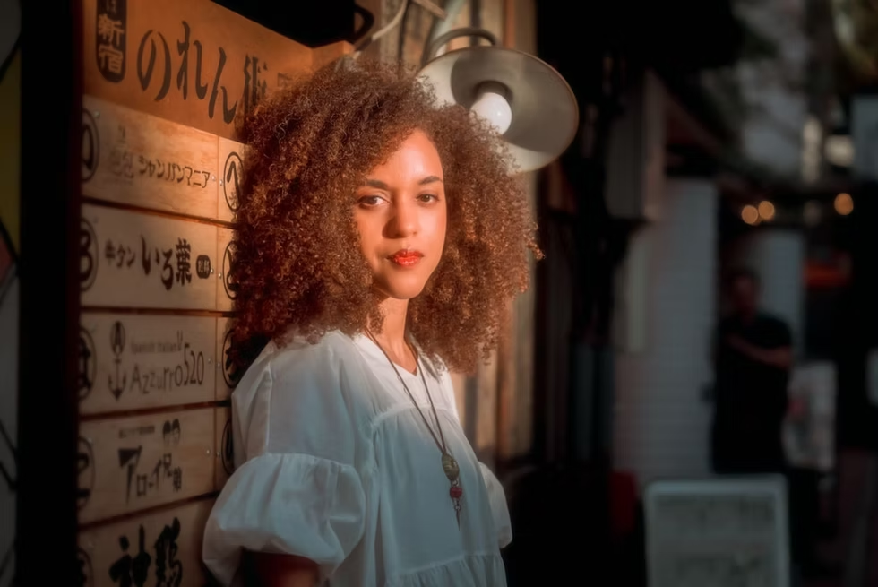 A woman with curly hair wears a white blouse and stands in front of a traditional Japanese restaurant sign. The background features dim lighting and Japanese characters on the sign.