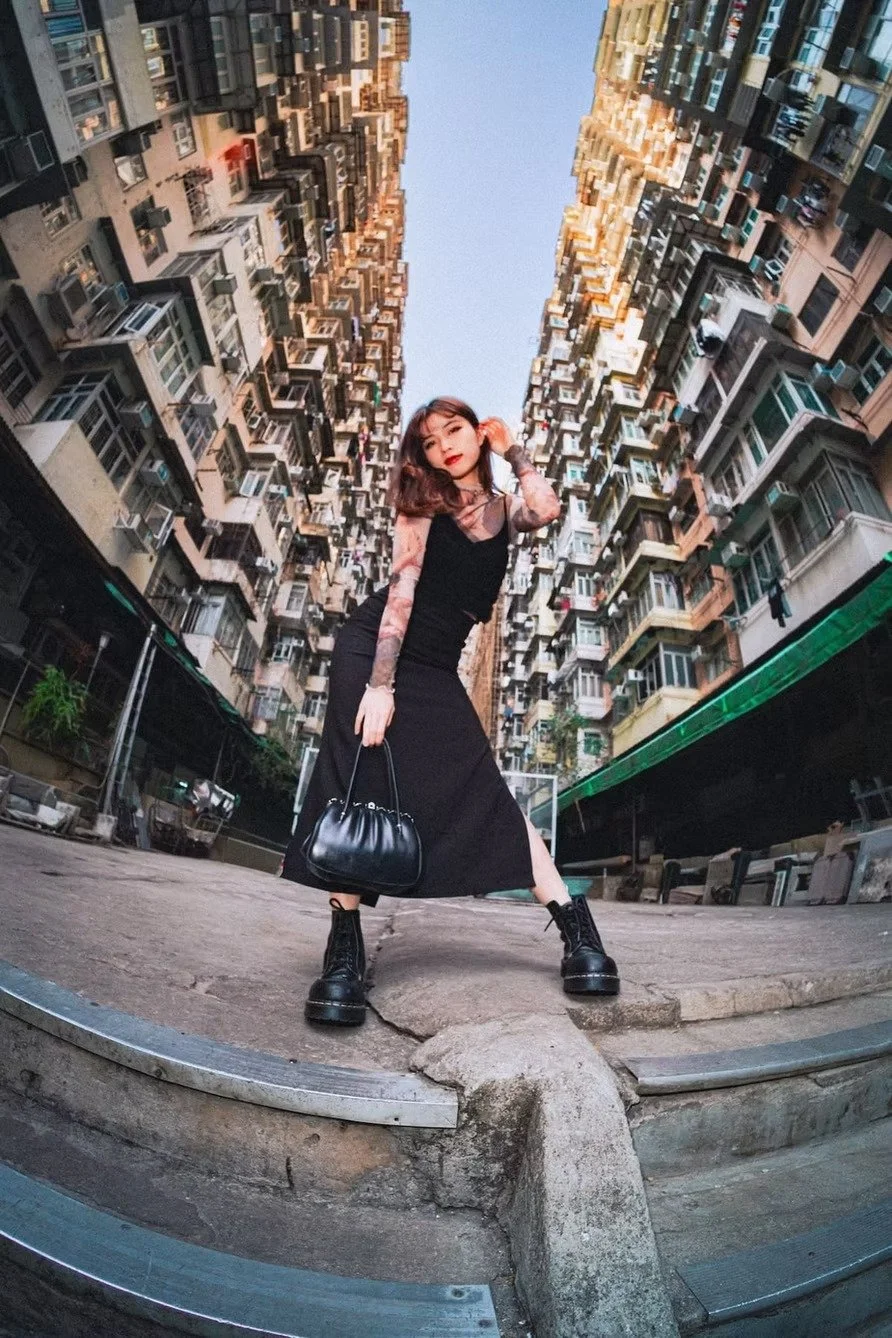 A young woman with shoulder-length hair poses in an urban alleyway between two tall apartment buildings, wearing a black dress, lace long-sleeve top, black chunky boots, and holding a black handbag, with a blue sky above shot at the Monster Building 