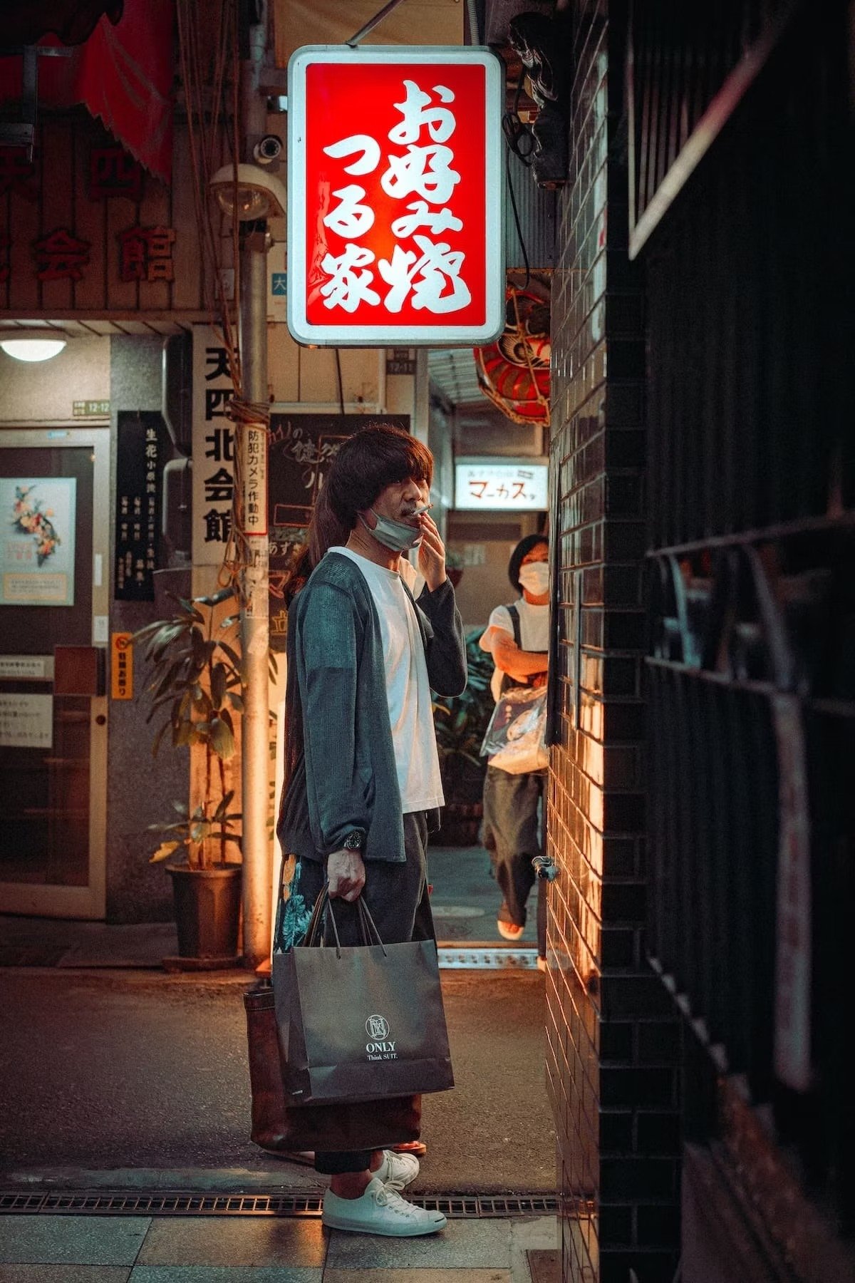 A man wearing a mask, hoodie, and sneakers holding shopping bags, standing on a city street at night, illuminated by a red and white sign with Japanese characters in Osaka.