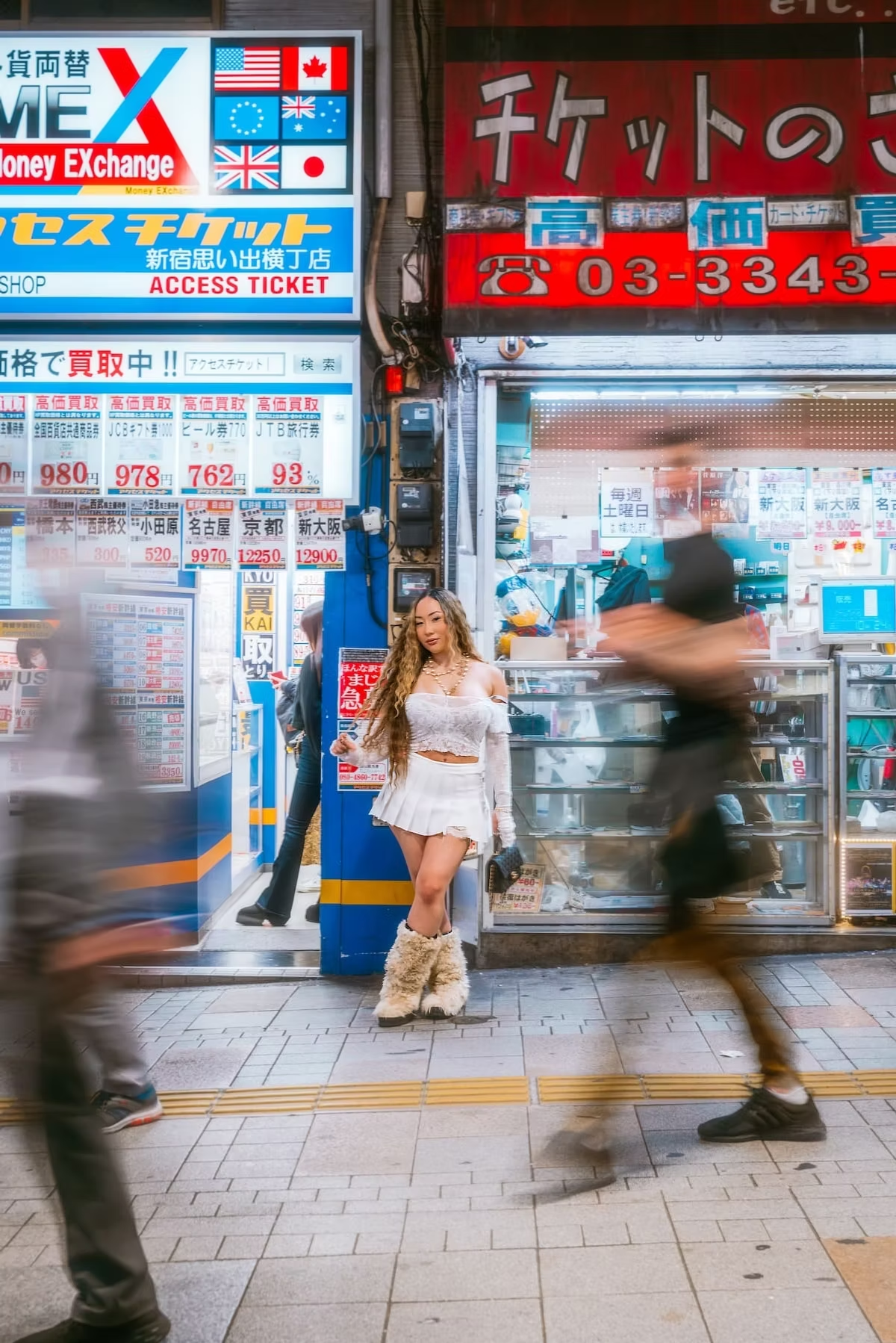 A woman in a white outfit with fur boots stands in front of a busy ticket shop in Tokyo. The area is crowded with blurred motion of people walking by, and neon signs light the scene.