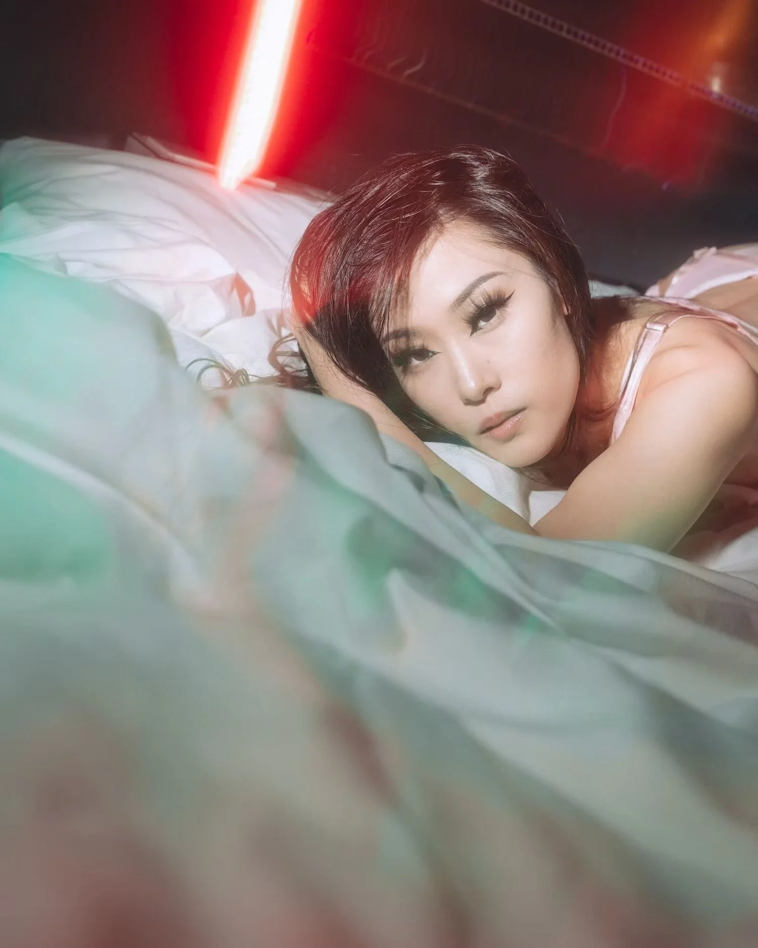 A woman with dark hair and makeup lying on a bed with white sheets, looking at the camera, with red and green lighting in the background.