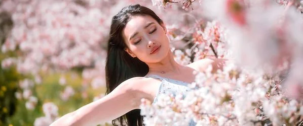 A woman in a white dress standing among pink cherry blossom trees with her eyes closed and arms open.