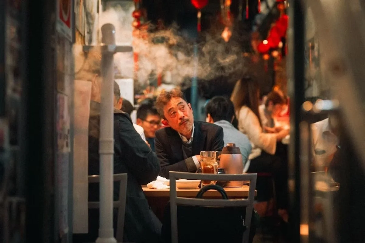 A Japanese man with gray hair and a beard sitting at a restaurant table, looking upward. There are several other people in the background, and the restaurant has a cozy, dimly lit ambiance with red decorations.