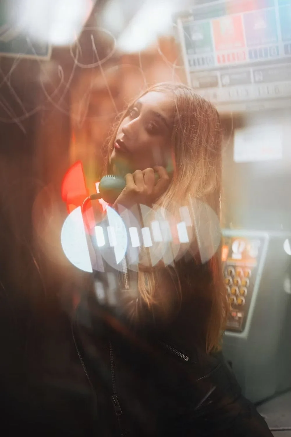 A woman with wavy hair talking on a vintage corded telephone inside a dimly lit room, possibly a recording studio, with various electronic equipment and a colorful sound mixer or control panel in the background.