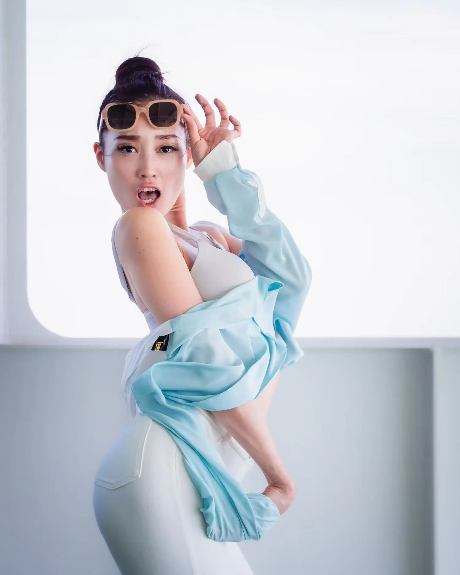 A young woman with dark hair in a bun, wearing sunglasses on her head, posing with her mouth open, and wearing a light blue garment.