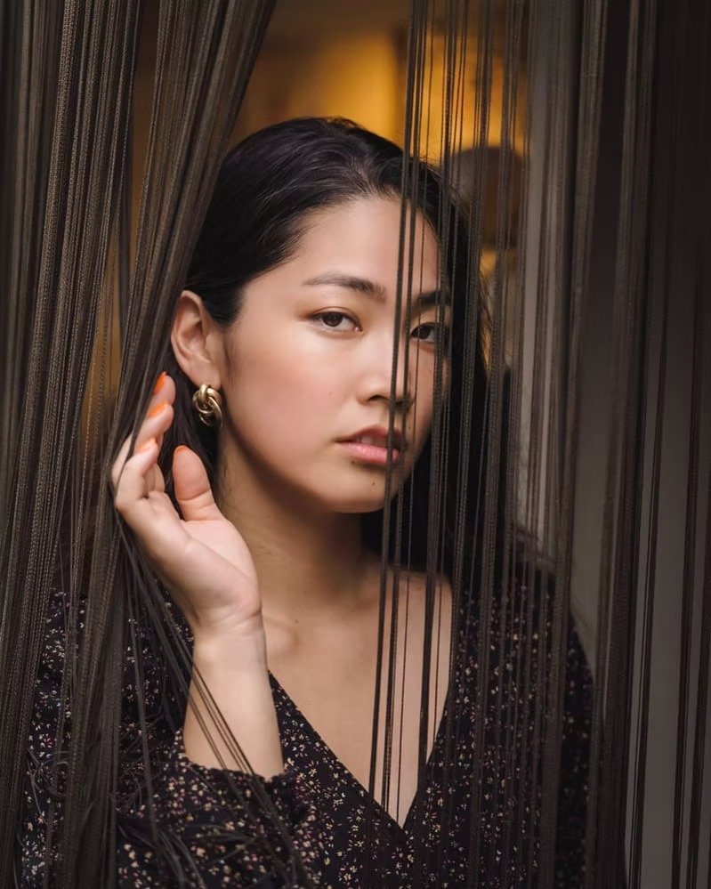 A young woman with dark hair and wearing a black dress, standing behind black sheer curtains, looking at the camera with a neutral expression.