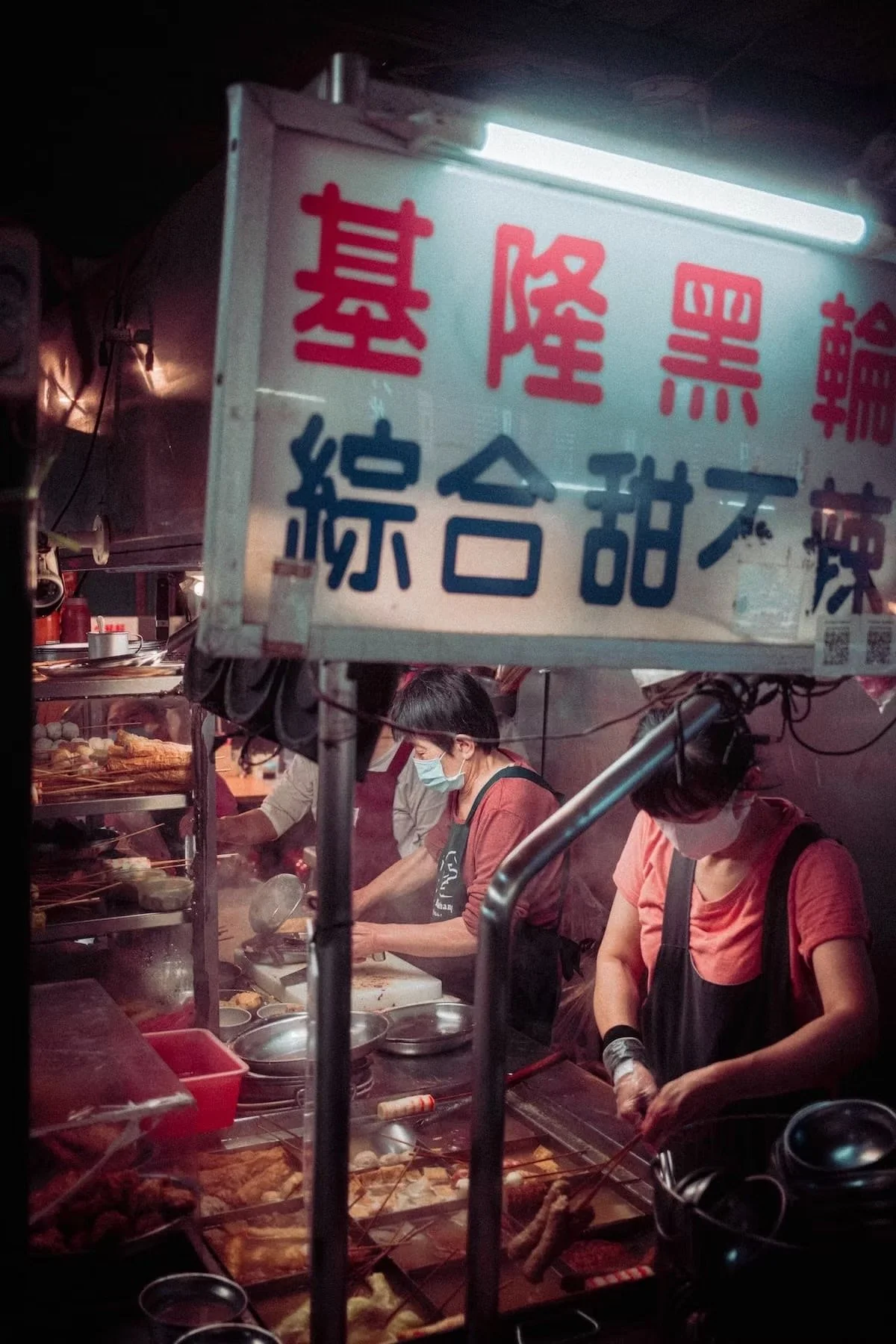 A street food stall at night with two women working behind the counter, wearing masks and black aprons, preparing food. The stall has a sign with Chinese characters and various food items are visible, including skewers and fried snacks. Taipei night 