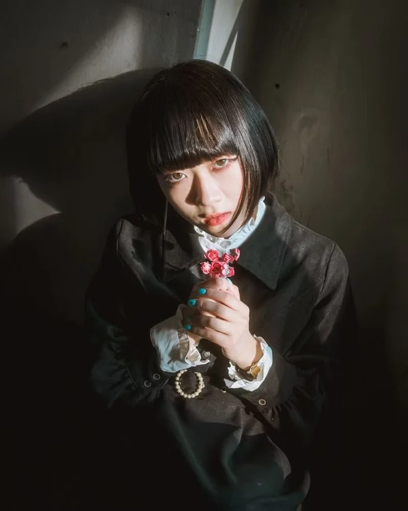A young woman with a chin-length black haircut, wearing a dark blazer and white shirt, holding pink flowers with both hands, looking directly at the camera with a shadowed background.
