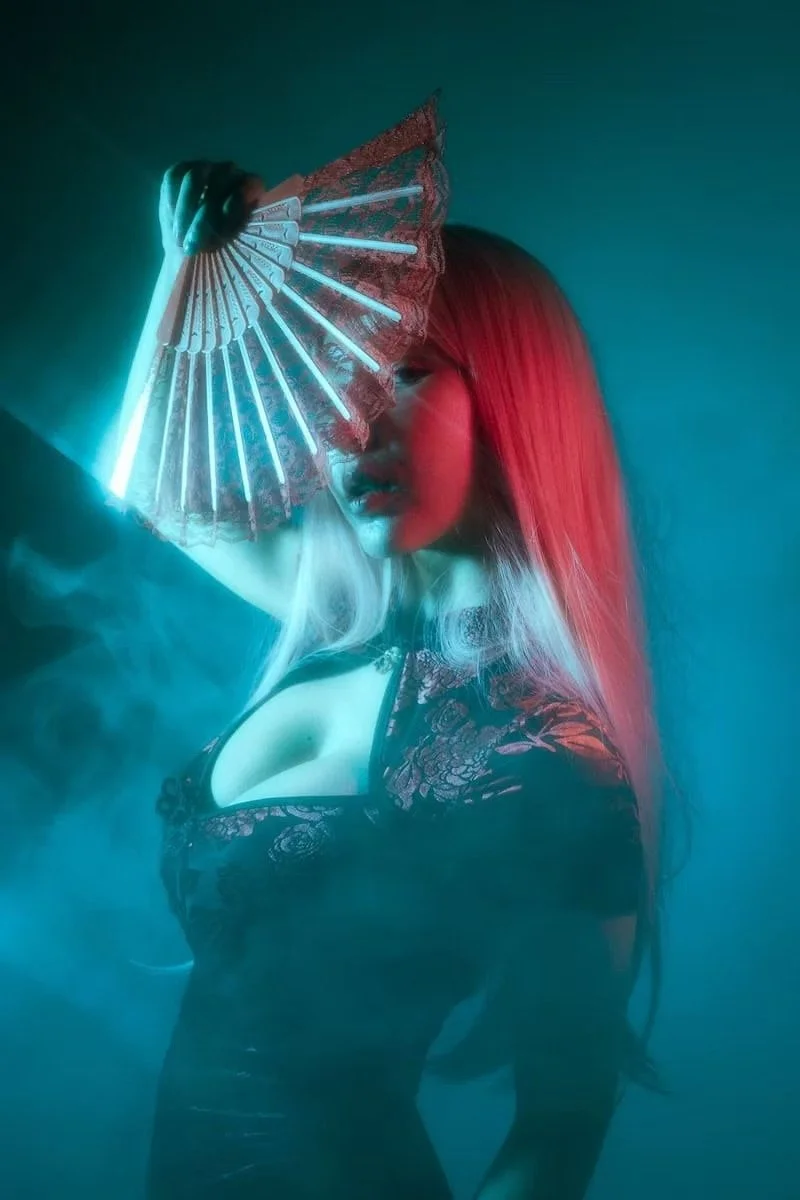 Woman with long hair holding a glowing, decorative fan in a dark, smoky setting with colorful lighting.