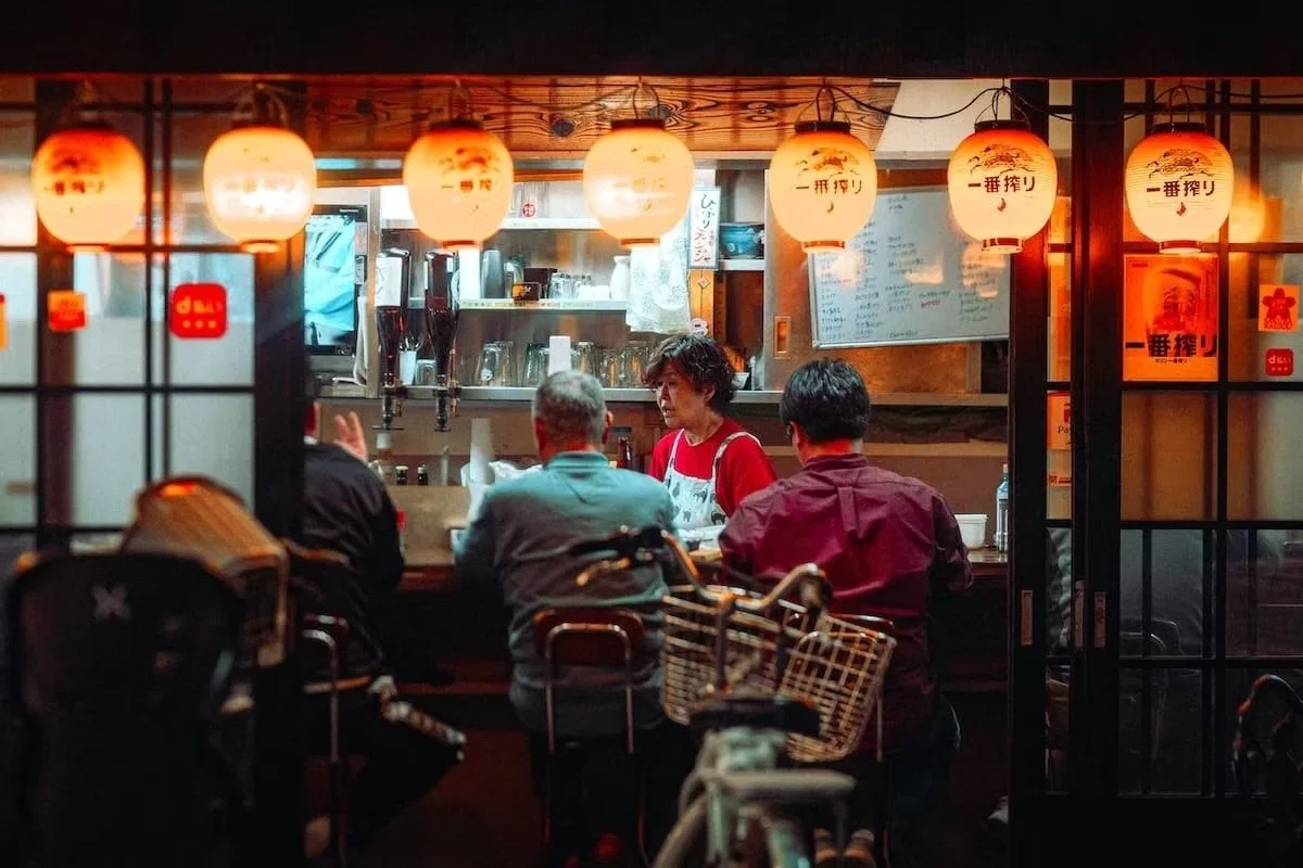 People dining at a small Japanese restaurant with paper lanterns hanging overhead, a chef behind the counter, and a menu on the wall in Osaka shot by Tokyo street photographer Deniz Demir.
