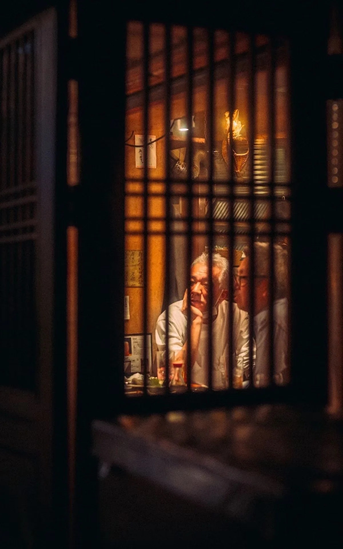 Two elderly men sitting at a table in a dimly lit traditional Japanese restaurant, seen through wooden lattice screens, with warm interior lighting and framed artwork on the wall in Ikebukuro Japan.