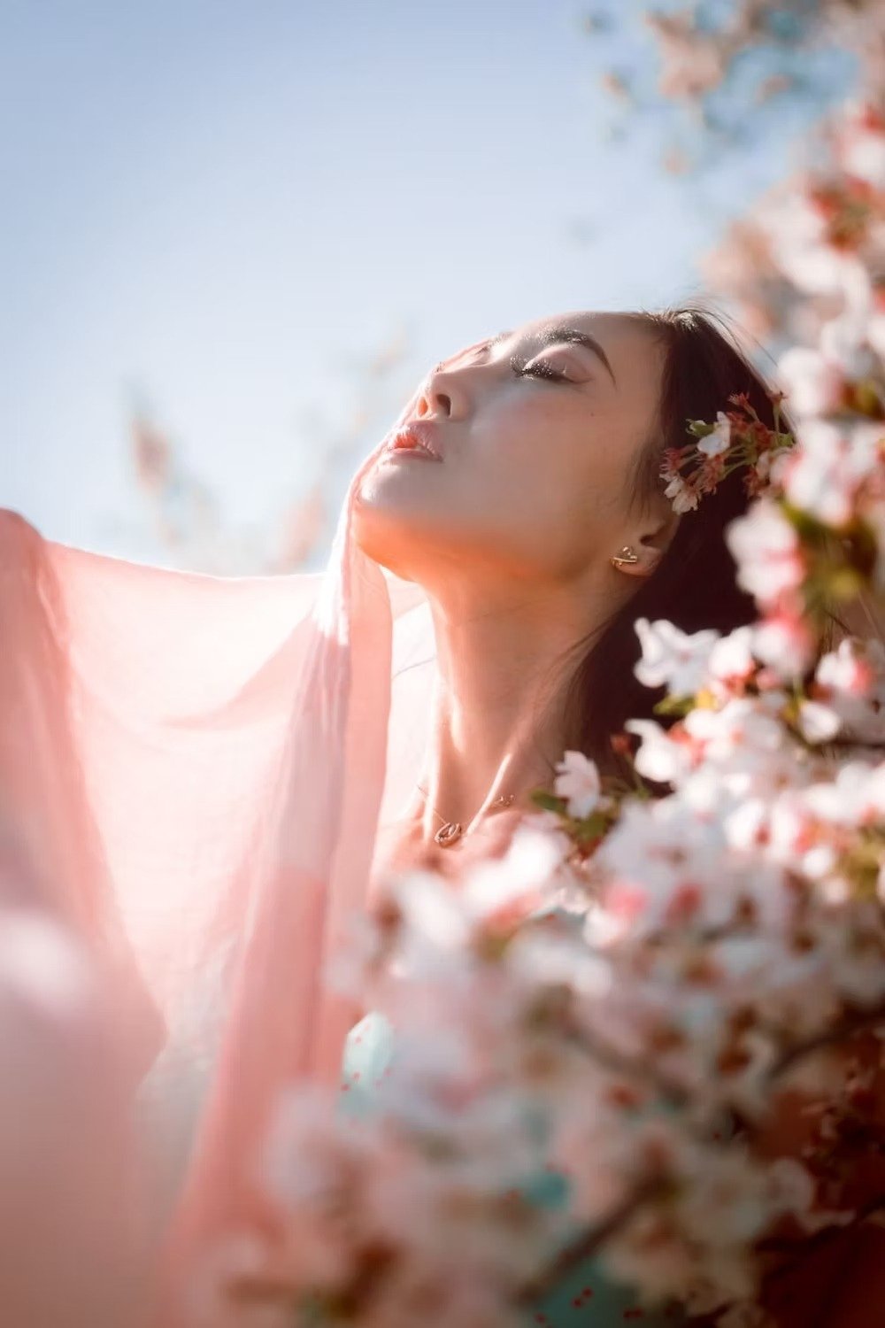 A woman with closed eyes and relaxed expression, surrounded by pink cherry blossoms, sunlight illuminating her face, in a serene outdoor setting. Portrait photography by Deniz Demir.