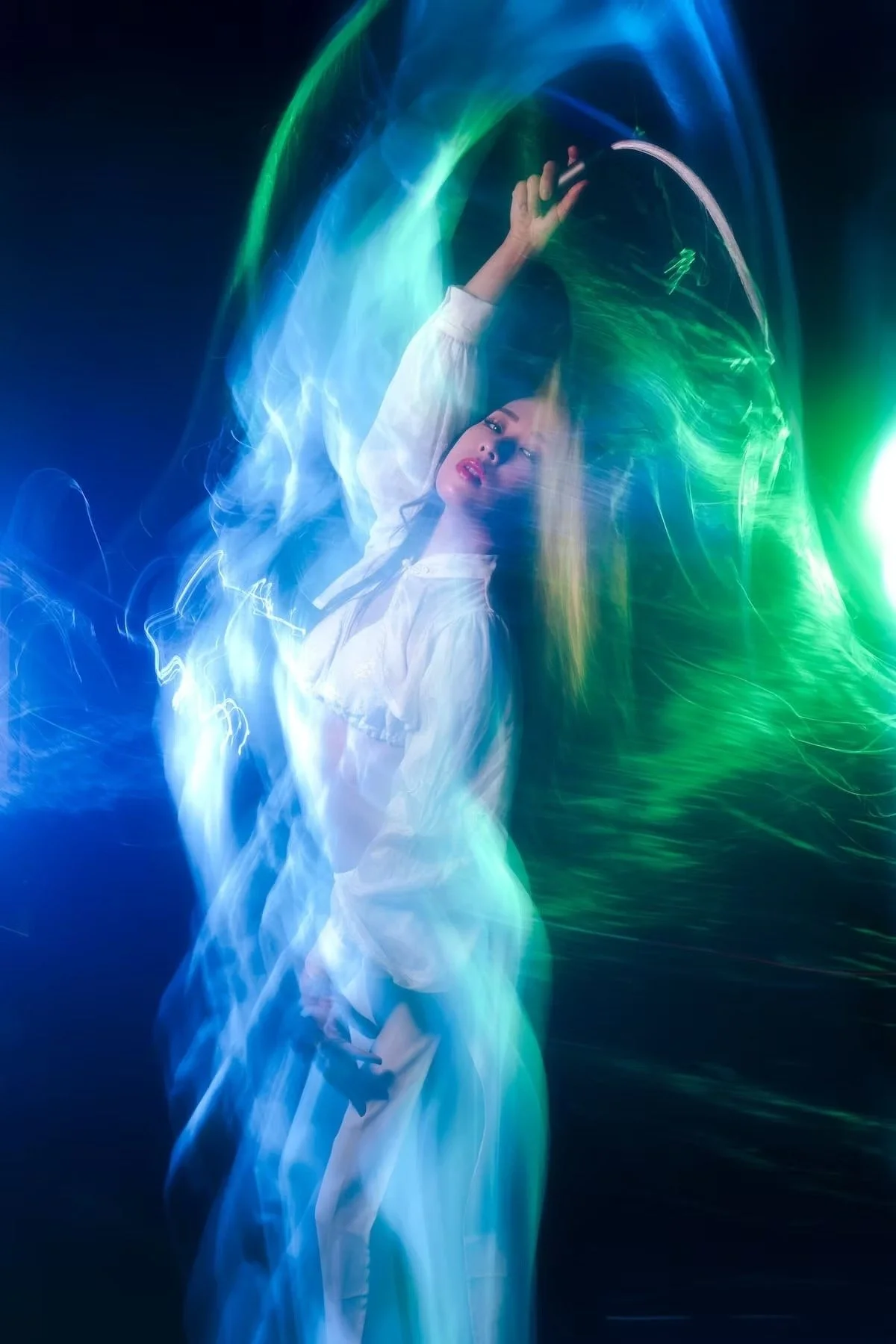 A woman with long hair, dressed in white, is lying on her back with her arm extended upward. She is surrounded by swirling, colorful lights in shades of blue, green, and purple against a dark background, creating a dreamy, abstract atmosphere.